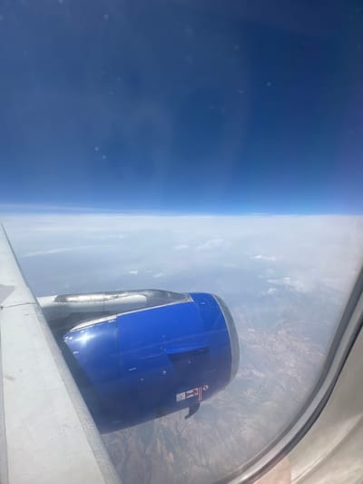 The image shows an aerial view from an airplane window. The foreground features the wing and engine of the aircraft, with the engine housing painted a distinctive blue. Beyond the aircraft, a layer of white clouds stretches across the horizon, separating the blue sky above from the land below. The land appears as a textured expanse of browns and greens, suggesting a rural or mountainous terrain. There are no identifiable structures or landmarks that would place this scene specifically in Viehhausen, Austria. The image captures a typical scene from a commercial flight, with a focus on the aircraft's mechanics and the vast expanse of the sky and clouds.