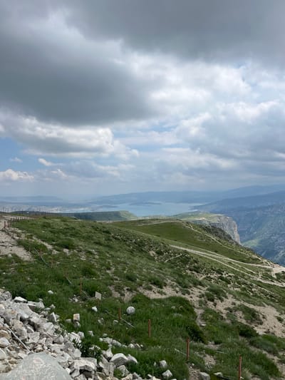 The image shows a view from a high vantage point, likely a mountain or steep hillside, overlooking a body of water and distant mountains. The foreground consists of rocky terrain transitioning into a grassy slope, with several small fence posts visible. A winding path can be seen traversing the hillside. The sky is cloudy, with a mix of light and dark cloud formations. The body of water stretches into the distance, separating the near hillside from the mountains on the opposite shore. The overall scene suggests a natural landscape setting. There is no evidence of people present in this image. Chirkey, Russia, is not discernable from this photograph.