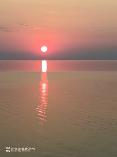 The image depicts a serene sunset over the Black Sea at Kamen Bryag, Bulgaria. The sun, a vibrant red-orange orb, is positioned low on the horizon, casting a warm glow across the sky and water. A bright, vertical reflection of the sun stretches across the calm sea, creating a striking visual. The water exhibits gentle ripples, suggesting a tranquil atmosphere. The sky transitions from a pale orange near the horizon to a muted gray at the top. A faint watermark in the bottom left corner reads "Shot on BL8800 Pro Powered by Blackview." There are no people or discernible objects present in the scene, only the natural elements of the sky and sea during sunset.