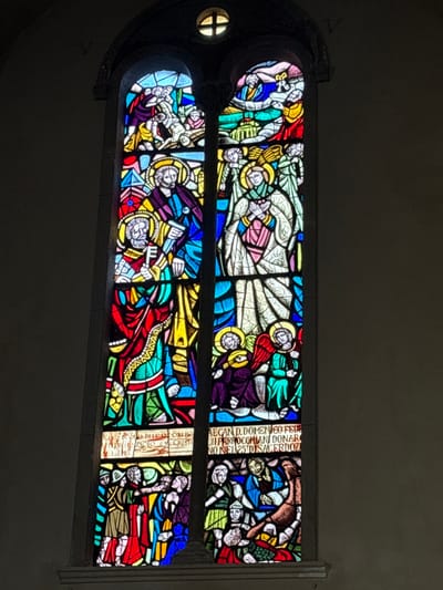 This image displays a tall, arched stained-glass window within a building located in the city of Foligno, Italy. The window is vertically divided into two main panels, with a smaller circular, multi-pane window featuring a cross motif positioned above. The surrounding wall is a plain, light gray.
The stained-glass panels are vibrantly colored in blues, reds, greens, yellows, purples, oranges, and whites, depicting various religious scenes.
**Left Vertical Panel:**
* **Top section:** A chaotic scene with multiple figures, some struggling or carrying a horizontal figure, set against a backdrop of hills and architectural elements. Colors are predominantly red, blue, and yellow.
* **Middle section:** Two standing male figures with halos are depicted. The figure on the left holds a large key and a book, while the figure on the right holds a book or tablet. Both wear layered robes in blues, reds, and gold.
* **Bottom section:** A group of figures, some in red robes, others in blue and green, are shown interacting. One figure appears to be gesturing or addressing others. An inscription below this section is partially legible, including the word "CRISTI."
**Right Vertical Panel:**
* **Top section:** A central figure looks upwards, surrounded by several smaller, winged angelic figures with halos, against a background of blue and green.
* **Middle section:** A tall, standing female figure in white robes with hands clasped in prayer is prominent, adorned with a halo. Below her, two smaller, seated angelic figures with halos and wings are depicted, one in purple and one in green.
* **Bottom section:** This section shows multiple figures engaged in various actions. Some figures are in blue and green robes; one figure appears to be falling or interacting with a brown, animal-like form. An inscription is present above this section, partially reading: "AEGAN D. DOMENICO FEDE," "IL PARROCCHIANI DONA," and "NO NEL 25 D'SACERDOZ."
Sunlight passes through the stained glass, illuminating the detailed figures and scenes with saturated colors, contrasting with the muted tone of the surrounding wall.