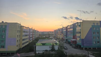 The image captures a view of apartment buildings in the city of Udachny, Russia, during what appears to be sunset. The sky exhibits a gradient of colors, transitioning from pale blue at the top to warm orange and pink hues near the horizon, with scattered clouds. Two long, multi-story apartment buildings, painted in vibrant geometric patterns of yellow, purple, green, and red, dominate the foreground and mid-ground, facing each other across a paved courtyard. These buildings have multiple floors, each with numerous windows and balconies, and are fitted with satellite dishes on their roofs.
In the courtyard between the buildings, several cars are parked, including a dark SUV, a silver minivan, and other vehicles. A smaller, single-story building with a patterned facade and an air conditioning unit is visible in the center of the courtyard. Trees and bushes are sparsely planted around the courtyard and along the sides of the buildings. In the background, more apartment buildings of similar design and scale extend into the distance, creating a densely built urban environment. Streetlights are visible in the foreground. The overall scene depicts a residential area in a northern climate, likely experiencing the extended daylight of summer or the transitional light of sunset. No people are clearly visible in the frame.