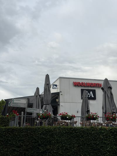 The image shows the exterior of a Woolworth store in Stadtallendorf, Germany. The store is a single-story building with a white facade. A large red "WOOLWORTH" sign is prominently displayed on the front of the building. Below the sign, there is a black and white sign with the letters "F&A". To the left of the building, there are several directional signs indicating routes to "Marktplatz," "Hatzbach," and "Haartsiedlung," as well as "Rathaus/Stadthalle" and "Bahnhof." The area in front of the store appears to be an outdoor seating area with tables and chairs, partially covered by large grey umbrellas. Flower boxes with red and white flowers are placed along the railing of the seating area. A dense green hedge runs along the bottom of the image, obscuring the lower part of the scene. The sky is overcast with grey clouds. There are no people visible in the image.