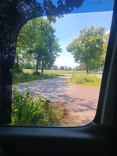 The image shows a view through a car window onto a rural road intersection on a sunny day. The road is paved with asphalt and gravel. To the left, there is a field of tall grass and bushes, with trees in the background. To the right, there is a gravel area and more grass. A yield sign with a bicycle symbol is visible at the intersection. Trees line the road in the distance, and a wind turbine can be seen in the far background. The sky is clear blue with some scattered clouds. The scene suggests a quiet, possibly agricultural area in or around Nordkirchen, Germany.