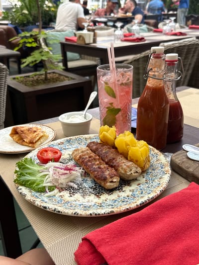 This image depicts an outdoor dining scene at a restaurant, likely in Zhukovka, Russia, given the context. The focus is on a table laden with food and drinks. In the foreground, a plate holds several grilled meat skewers, some boiled potatoes, a small portion of salad with sliced red onion and tomato, and a piece of folded pastry on a separate small plate. A small bowl of white dip, possibly yogurt or sour cream, is also visible with a spoon. To the left, a tall glass contains a pink beverage with ice and mint leaves, served with two straws. Behind the drinks, two glass bottles with metal swing tops contain reddish liquids, likely sauces or condiments.

In the background, the scene suggests a bustling restaurant environment. Several people are visible, some seated at tables, with one person wearing a white shirt and another in blue attire. The setting appears to be a patio or terrace, with woven chairs and tables, and greenery in planters, hinting at a pleasant, possibly warm, time of day. The overall ambiance suggests a relaxed meal enjoyed outdoors. The time of day is likely daytime or early evening, given the lighting. No text is visible in the image.