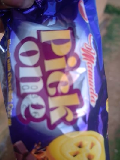 The image shows a close-up of a partially opened package of cookies, likely "Pick Up" brand, with the prominent logo in purple and yellow. The packaging is shiny and reflective, with some text visible on the left side in a smaller font. The cookies themselves appear to be chocolate-filled or chocolate-coated, with a textured surface visible at the bottom of the image. The background is blurred and appears to be outdoors with some greenery and a dirt or paved surface. There is no discernible indication of the specific location within Rahwol Chom, Nigeria, nor are there any people or specific actions depicted in the image.
