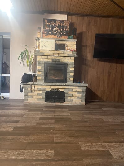 The image depicts an indoor domestic space, likely a living room, in Petrivka, Ukraine, characterized by a central brick fireplace, a mix of wall finishes, and dark wood flooring. **Environment:** The floor is covered with dark brown, wood-patterned tiles arranged in a staggered horizontal pattern, simulating distressed wood planks. The left wall is painted a light pinkish-beige with a subtle vertical striped texture, while the right wall and ceiling are clad in dark brown wooden paneling, with the ceiling featuring horizontal slats. A darker wooden beam runs along the junction of the ceiling and the right wall. An open doorway on the far left reveals an adjacent room; two white slippers are on the floor near its threshold. **Central Objects:** A prominent fireplace, constructed with alternating light beige and dark gray rectangular bricks, serves as the focal point. It features a light gray, granite-like mantle and hearth. A black metal, glass-fronted fireplace insert is embedded in the brickwork, displaying active flames or embers within. Below the main firebox, a decorative black metal grate covers an opening, likely an ash pit or storage area. **Objects on and Around the Fireplace:** Above the fireplace, on the light-colored wall, a large framed photograph depicts a group of approximately ten to twelve individuals, primarily men, dressed in dark attire. On the upper fireplace mantle (shelf), a tall green houseplant in a white pot is situated on the left, alongside a black backpack and a brown shoulder bag. On the right side of the mantle, a small clear glass jar, a blue object (possibly a lighter), and part of a string light are visible. The lower mantle (hearth) displays a framed picture with a circular bamboo motif. To its left are two bottles: one labeled "LIBERTIES The Gin" and another, partially visible, labeled "AZNAURI." To the right of the framed picture, a small gold frame contains four green circular objects, and a collection of miniature liquor bottles rests on a small tray. **Other Features:** On the right wood-paneled wall, a large, dark flat-screen television is mounted and is currently off; associated cables are visible beneath it. The room is illuminated by ambient light, with a strong glare emanating from an unseen light source in the upper left corner, possibly from a window or a bright overhead fixture. **People/Actions:** No individuals are distinctly visible in the immediate foreground. A blurred, indistinct figure or reflection of a
