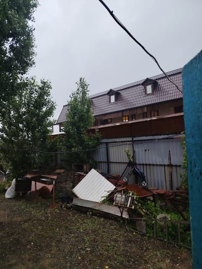 This is a moderately distant, eye-level, medium shot of an outdoor, cluttered backyard area with a two-story building in the background under an overcast sky. The building has a dark, tiled roof with two dormer windows. A corrugated metal fence runs horizontally across the middle ground, separating the yard from the building. The yard is filled with various objects, including stacks of tiles or roofing material, a metal grill, a white garbage bag, a dark bucket, a disassembled stroller, and a large white angled piece of material, possibly a shed roof or siding. Green foliage is present, with trees to the left of the building and vines growing on the fence and near the pile of debris. The ground is covered in gravel and dirt, appearing wet, suggesting recent rain. The overall impression is one of disarray and accumulated materials. The location is stated to be in Sol-Iletsk, Russia, which is a city in Orenburg Oblast known for its salt lakes and spa resorts.