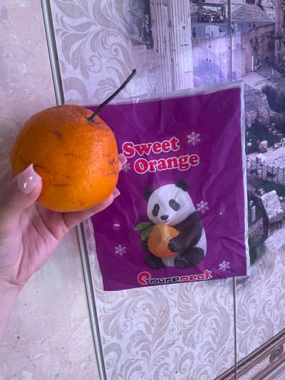 A hand with pale pink fingernails holds an orange with a dark twig protruding from its top. The orange is rough-skinned and has some dark blemishes on its surface. Behind the orange, a purple paper with a printed image of a panda holding a piece of orange and the words "Sweet Orange" and "purepeak" is visible. The background consists of a tiled surface with a floral pattern and a larger image of what appears to be a cityscape or architecture. The overall scene suggests a casual indoor setting. The lighting is somewhat dim, making it difficult to determine the time of day or weather conditions. There is no clear indication of a specific event taking place, other than the presentation of the fruit and packaging. The colors are muted, with the purple paper and orange fruit being the most vibrant elements. The focus is on the orange and the packaging, implying they are the subjects of interest.