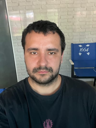 A mid-shot, eye-level, front-facing selfie shows a man with dark, wavy hair, a mustache, and stubble. He is wearing a black t-shirt. Behind him, to the right, is a blue folding chair with "1664" printed in white on the backrest. The background consists of a light-colored brick wall. The scene appears to be indoors or on a covered patio, and there is no indication of the city of Nikopol, Ukraine, or any other specific location.