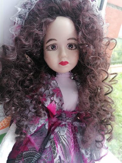 The image shows a close-up of a porcelain doll with long, dark, curly hair and dramatic makeup. The doll is wearing a layered dress with a pink, purple, and gray patterned fabric and a delicate white lace trim on its head. The doll's face is light-skinned with bright brown eyes, dark eyebrows, and red lips. The background is blurred, suggesting an outdoor environment with greenery visible. The scene does not depict any specific location within the city of Starye Igiti, Russia.