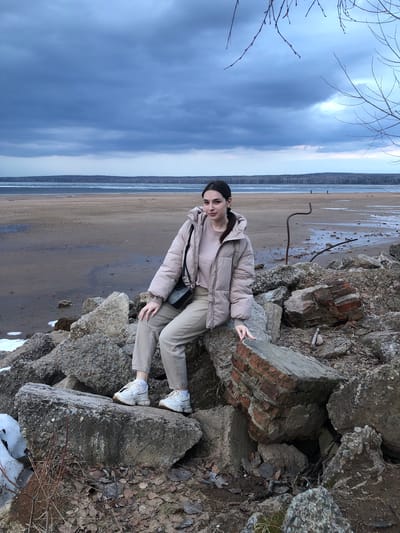 A young woman stands among the ruins of what appears to be a brick and concrete structure. She is wearing a light pink puffer jacket and khaki pants with white shoes. Behind the ruins is a wide expanse of water and sand, possibly a beach or a riverbank, under a cloudy sky. The location is the city of Kizhi, Russia.