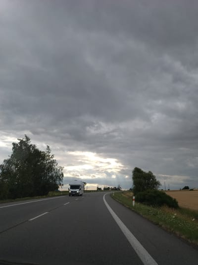 The image depicts a two-lane asphalt road under a dramatic, cloudy sky. A white camper van is traveling on the right lane of the road, moving away from the viewer. On the left side of the road, there is a dense, leafy green tree. The right side of the road is bordered by a field of dry, golden crops and green vegetation with scattered white wildflowers. Several white posts with red reflectors are visible along the side of the road. The sky is filled with heavy, dark gray clouds, but shafts of sunlight break through in the distance, illuminating the horizon. The overall impression is that of a rural landscape on an overcast day, possibly during late afternoon or early evening. The location is identified as a road in the vicinity of Tábor, Czechia.