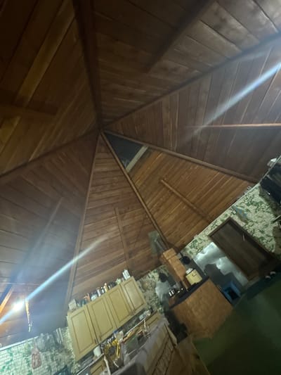 The image depicts the interior of a wooden structure with a pitched roof. The walls appear to be constructed of brick and are covered in a green and white pattern. In the center of the scene, there is a brick oven or fireplace. To the right, a wooden door is visible. On the left side of the image, there are wooden cabinets with items placed on top. The lighting suggests it is evening or nighttime. No individuals are present in the frame. The scene is likely located in the city of Yabogan, Russia, given the provided context.
