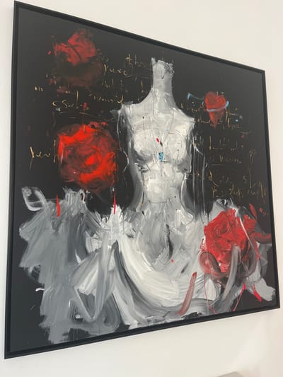 This image depicts an abstract expressionist painting, mounted on a plain white wall, located within a building in Galatina, Italy. The painting features a dark, predominantly black background, overlaid with broad, visible brushstrokes. A central, headless, and armless dressmaker's mannequin or dress form is rendered in white and gray tones, occupying the central and lower-mid sections of the canvas, depicted with an implied garment. A small blue pendant or detail is visible near the chest area of the mannequin. Three abstract, textured red forms, resembling roses or floral elements, are prominently placed: one in the upper left quadrant, one in the mid-left, and one in the lower right. Illegible script or scribbled markings in a golden or yellow hue are scattered across the dark background. The artwork is enclosed within a thin, black frame.
