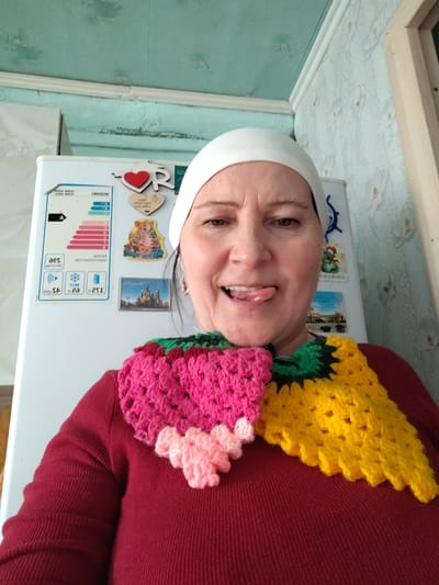 A medium close-up selfie shows a woman indoors, possibly in a kitchen or home setting. She is looking directly at the camera with a playful expression, her tongue extended. She wears a white head covering, resembling a beanie or underscarf, and a dark red long-sleeved sweater. A distinctive, multi-colored crocheted neck warmer or scarf is draped around her neck and shoulders. This item features sections of bright pink, light pink, green, and yellow yarn, arranged in a textured, block-like pattern, possibly depicting a fruit like a strawberry or pineapple.
In the background, a white refrigerator is visible, covered with various magnets. These include a red heart with a white "R", a wooden heart magnet with the word "wedding" written on it, a smiling tiger, a snowman, and small magnets depicting architectural scenes. A prominent energy efficiency label is affixed to the refrigerator door, displaying an "A" rating and other specifications. The text on this label appears mirrored, showing phrases like "SUSUS" (likely reversed "SAMSUNG") and "ЗНЭИУЕПЬО" (likely reversed Cyrillic for "energy efficiency").
The surrounding environment features light blue wallpaper with a subtle pattern and a light blue ceiling. The overall impression is casual and personal, captured in what appears to be a home in Сусады-Эбалак, Russia.