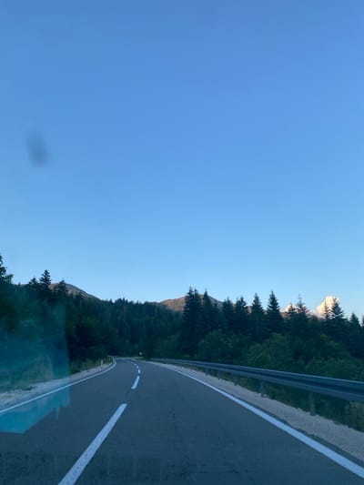 An image taken from a moving vehicle shows a winding asphalt road through a forest of coniferous trees, with a clear blue sky above. The road has white lane markings. A metal guardrail runs along the right side of the road. In the distance, mountains are visible, partially obscured by trees. There is a faint reflection on the left side of the image, likely from the vehicle's window. There are no people or animals visible. The scene appears to be in a mountainous, natural environment. The location is described as the city of Praga, Montenegro.