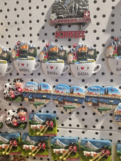 Here is a detailed description of the image:

The image shows a display of souvenir magnets likely for sale. The magnets are arranged on a white board with evenly spaced holes. Several different designs are visible, all related to Switzerland. These include:

*   A silver-colored magnet with a landscape of Switzerland and the words "SWITZERLAND" and "SCHWEITZ".
*   Magnets shaped like a cup with half of a Swiss townscape on one side and the words "I [heart] BASEL" printed on the other.
*   Heart-shaped magnets with a view of Basel, Switzerland, including the Rhine River.
*   Magnets depicting Swiss cows.
*   Magnets showing Swiss Alpine scenes with individuals playing an alpenhorn.

The image does not show any specific location in the city of Saint-Louis, France. It only depicts a display of magnets.