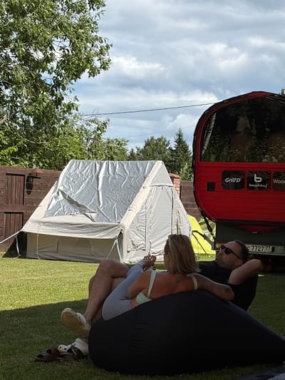 In Shebanovo, Russia, a man and a woman relax on a large black beanbag chair outdoors on a sunny day. The woman, facing away from the camera, is wearing a light green top and light gray leggings. The man, wearing a black t-shirt and sunglasses, is lying down with his head propped up. Behind them, a large, light-colored tent with a silver cover is set up on a grassy lawn. To the right of the tent is a distinctive red structure, possibly a mobile sauna or vehicle, with the logos "Grill'D", "bochky", and "Wood" visible on its side. A license plate with the numbers "C 7727 77 RUS" is affixed to the rear of the red structure. A wooden fence borders the left side of the scene, and a tree with green foliage is visible in the background, under a partly cloudy sky.