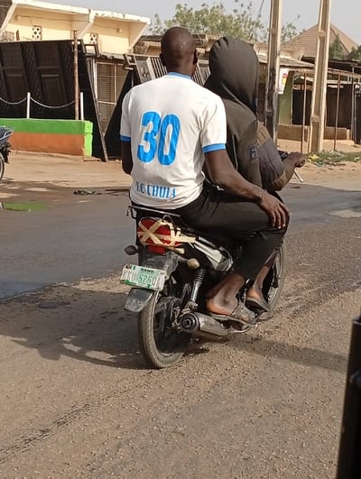 Two males are observed from behind, riding a dark-colored motorcycle on a light brown, unpaved road in Gayawa, Nigeria. The driver, with a bald head and dark skin, wears a white short-sleeved t-shirt featuring a large blue number "30" and "J.C. CHULA" printed below it, along with black trousers and sandals. His left arm is extended forward, presumably operating the motorcycle. Seated directly behind him is a passenger, clad in a dark grey hooded jacket with the hood up, black trousers, and sandals. The passenger's right hand rests on the driver's left hip. The motorcycle has a visible Nigerian license plate from Kano state, reading "01I KANO KNU 525QA", and a reddish-brown patterned item is strapped to its rear seat. The background features several light-colored buildings with dark metal doors and gates, some exhibiting green and orange painted sections on their lower walls. Sparse vegetation and dry trees are visible further in the background under bright daylight, casting shadows on the road surface.