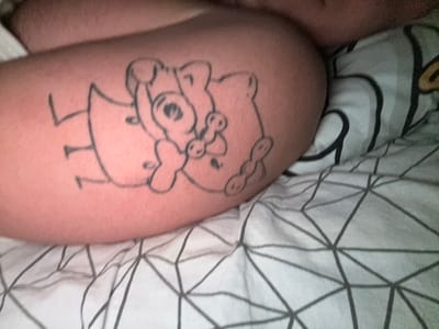 The image shows a tattoo on a person's leg or thigh. The tattoo depicts a cartoon-like drawing of two figures. One figure has a bow in its hair and is holding a pacifier. The second figure is partially obscured but appears to be wearing a skirt or dress. The background includes a bedsheet with a geometric pattern and a white pillow with a cartoon design.