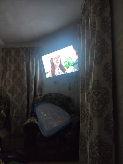 The image depicts an indoor scene, likely a living room, with dim lighting. A television screen displays a close-up of a woman's face. The television is mounted on the wall, angled downwards. To the left and right of the television are curtains with a damask pattern. In the lower part of the image, a plush armchair is visible, partially obscured by a blue mesh laundry basket containing items. The overall environment suggests a residential setting. The location is specified as Shaldai rural district, Kazakhstan.