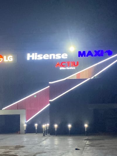 Illuminated signs for LG, Hisense, ACTIU (Office Furniture), and MAXI are stacked vertically on a building facade. The signage leads to a building with a striped facade highlighted by LED strips. A row of streetlights illuminates the adjacent pavement. The scene is captured at night, with a dark sky in the background.