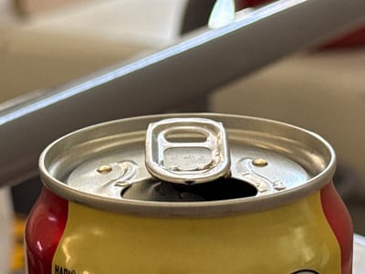 The image shows a close-up, eye-level view of the top of an open aluminum can with a red and yellow design. The can is sitting on a surface, and a metallic bar or rail is visible in the blurred background, angled downwards from left to right. The can's pull tab is raised, and a small droplet of liquid is visible on the silver lid near the tab. The can appears to be a beverage container. The scene is not identifiable as being in Troitske, Ukraine, or any specific city.