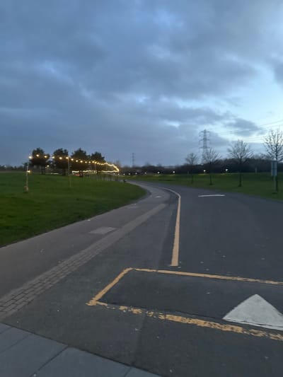 The image captures an outdoor park or recreational area in Falkirk, United Kingdom, consistent with the pathways and landscape of The Helix Park, during dusk or early evening under an overcast sky. No individuals are present.
In the foreground, a wide asphalt path or roadway curves from the lower right towards the mid-ground left. It features a continuous yellow line marking its center and a yellow rectangular box painted on the surface, within which an inverted white triangle (likely a yield or directional arrow) is visible. Adjacent and to the left, a separate, narrower paved pathway, partially surfaced with reddish brick pavers and partially asphalt, parallels the main path before curving left into the distance. Both paths are bordered by expansive, manicured green grass verges.
Along the left grassy verge, a string of warm yellow decorative lights is strung between a series of upright poles or slender trees, extending in an arc into the mid-ground. Further along this side, a cluster of trees with visible foliage is present. To the right of the main path, a sparse line of bare-branched deciduous trees stands. In the distant background, several tall metal lattice transmission towers (pylons) are silhouetted against the dark, cloudy sky, indicating a power line route. Additional individual streetlights or park lights are visible illuminating the path further in the distance. The sky is uniformly grey-blue with substantial cloud cover.