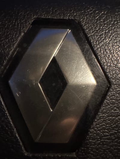 A close-up, top-down view shows a metallic Renault logo set into a dark, textured material consistent with pebbled leather or automotive upholstery. The logo features a hexagonal perimeter encompassing a central diamond design formed by two interlocking 'V' shapes. The metallic surfaces of the logo, which appear silver or chrome, exhibit numerous fine scratches across their reflective finish. The surrounding material is uniformly textured. No individuals, actions, or other distinct objects are present in the frame. The scene is located in the city of Sarcedo, Italy.