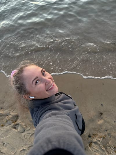 A woman with blonde hair pulled back and a pink scrunchie is standing on a sandy beach in Teriberka, Russia, near the water. She is wearing a grey fleece jacket and white earbuds. The water is calm with small waves lapping at the shore. Her body is angled away from the camera, and she is looking back over her shoulder with a smile. There are footprints in the sand around her.