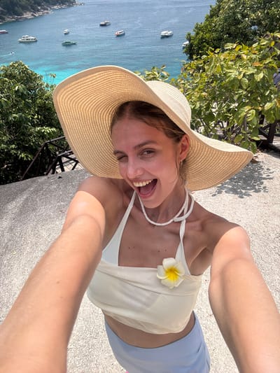 A young Caucasian woman is taking a selfie outdoors. She is wearing a large straw hat, a light-colored top with a white flower pinned to it, and light blue bottoms. She is smiling broadly with her mouth open. The background features a blue body of water with boats, lush green vegetation, and a gray concrete surface. The location is the city of Lam Kaen, Thailand.