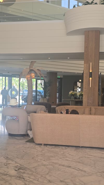 The image captures a brightly lit, modern indoor space with a resort-like ambiance, possibly a hotel lobby or lounge, situated in Parekklisia Community, Cyprus. The scene features a large, U-shaped, beige upholstered sofa with textured fabric, positioned on a marble-tiled floor. A person is seated on the sofa, facing away from the camera, engrossed in their mobile phone.
Further into the space, a distinctive decorative palm tree with woven fronds and exposed bulbs serves as a focal point, casting a warm glow. Behind it, large glass doors reveal an outdoor view of palm trees and what appears to be a street or parking area with vehicles, suggesting a warm climate.
The architecture is characterized by clean lines and natural materials, with a prominent wooden beam extending across the ceiling and white, ribbed wall paneling. There are several accent lights and a flat-screen television mounted on the wooden beam. The overall atmosphere is relaxed and contemporary. The lighting suggests it is daytime. No visible text is present.