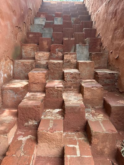 The image shows a set of rustic, weathered stairs constructed from red bricks and concrete blocks, nestled within a reddish-brown earthen structure that appears to be a wall or embankment. The stairs ascend upwards and to the right, with some steps being solid brick, others concrete, and some a combination. The surfaces are uneven and show signs of age and erosion. The overall color palette is dominated by earthy reds and grays. The setting is likely outdoors, given the natural lighting and the texture of the materials. This scene is located in San Agustín Etla, Mexico, specifically within the ruins of the former textile factory, now an art center.