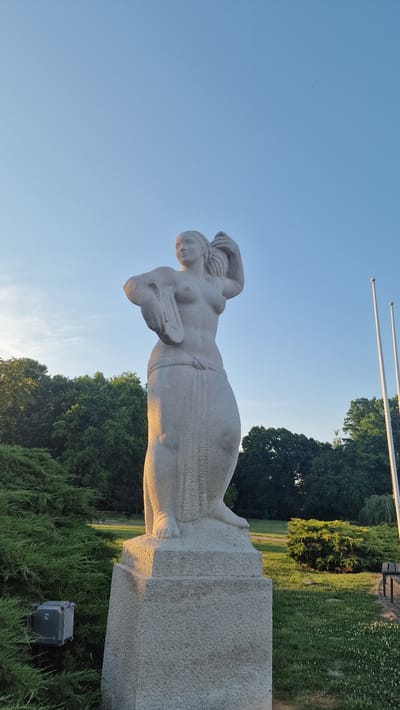 A white stone statue of a woman stands on a pedestal in a park. The statue is approximately life-size and depicts a nude figure with her hair pulled up, holding a piece of fabric in her left arm and a strand of hair in her right hand. She is wearing a draped cloth around her waist. The statue is illuminated by the golden light of a low sun, casting shadows on its surface.
The statue is located in Keszthely, Hungary, specifically in the vicinity of Balaton Museum. The background features lush green trees and a clear blue sky. There are two flagpoles visible to the right of the statue. In the foreground, a manicured evergreen bush is to the left of the pedestal, and a small electrical box is mounted on it. To the right, a grassy area with scattered white flowers and a park bench can be seen. The overall atmosphere is serene and picturesque, suggesting a tranquil outdoor setting.