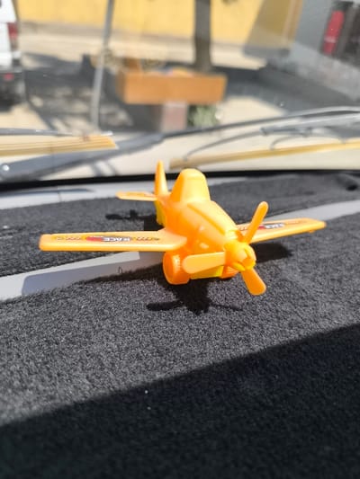 The image shows a close-up, eye-level view of an orange toy airplane with black and red detailing on its wings, resting on a dark grey, textured surface, likely a dashboard or floor mat. The airplane is positioned slightly to the right of center and is facing left, with its propeller slightly angled. Sunlight casts a shadow from the airplane onto the surface. In the blurred background, reflections on a glass surface (likely a car windshield) show a yellow structure, possibly a building or a cart, and hints of a vehicle. The overall environment suggests an outdoor setting, possibly within a vehicle. The location is identified as Kumbasti, Uzbekistan.