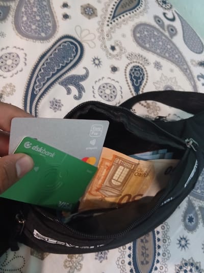 The image shows a partially opened black fanny pack resting on a surface with a paisley pattern in shades of white, grey, and blue. Inside the pack, there are euro banknotes. In front of the fanny pack, a hand holds two credit/debit cards, one silver ("EasyPay" logo is visible), and one green ("dskbank" logo is visible). The location is presumed to be Vetren Dol, Bulgaria.
