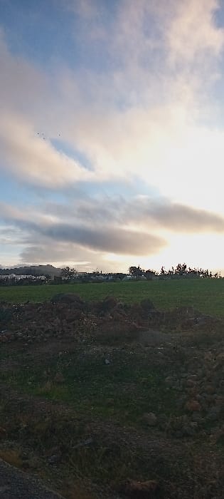 The image depicts a rural landscape in Ait Naamane, Morocco. The scene is dominated by a partially cloudy sky, transitioning from light blue in the upper left to a bright, sun-illuminated cloud formation on the right, suggesting either sunrise or sunset. Five small, dark avian silhouettes are visible flying in the upper left quadrant. The midground features a large, level green field. Beyond this field, a distant village or settlement with scattered buildings and trees is observable, backed by a low-lying hill range on the far left. The immediate foreground consists of uneven, uncultivated terrain composed primarily of dark earth and numerous brown rocks, with sporadic patches of sparse green vegetation. No individuals, specific actions, or interactions are discernible within the frame.