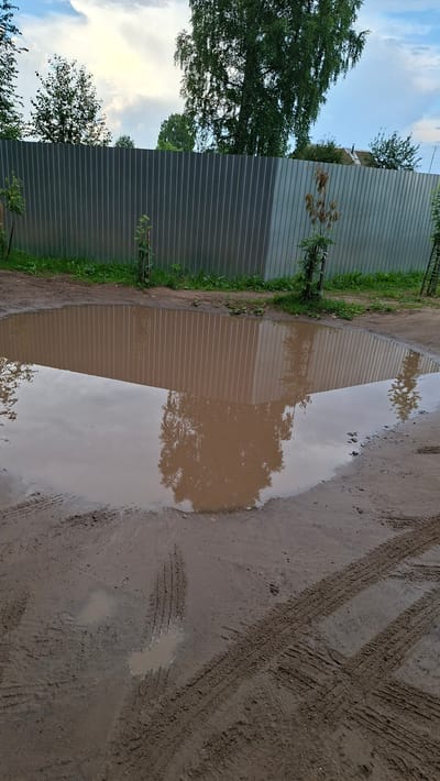 This is a ground-level, vertical shot of a muddy area in Novoilinsky, Russia, on a cloudy day. A large, shallow puddle of murky brown water dominates the center of the frame, reflecting the grey corrugated metal fence that stretches across the background. The fence is backed by a strip of green grass and vegetation, with trees and a building visible beyond. The foreground consists of wet, disturbed earth with prominent tire tracks imprinted in the mud. There are no people or animals visible in the image.