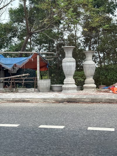The image depicts a roadside scene in Quảng Trị Ward, Vietnam. In the foreground, a wet asphalt road is visible with white dashed lines indicating lanes. Beyond the road, a concrete curb separates the road from a paved area where several large, grey concrete vases are displayed. Two prominent vases, tall and ornate, stand in the right portion of the image. To the left of these vases, a makeshift shelter is set up, covered by a blue tarp and an orange tarp. Beneath the shelter, wooden structures are visible, along with various concrete planters and what appear to be ceramic pots. Behind the shelter and vases, lush green trees and foliage form a dense backdrop. The overall impression is of a small roadside stall selling decorative concrete items. There are no people visible in the image.
