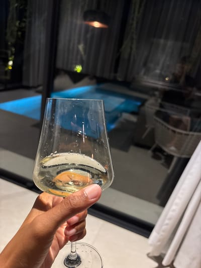 The scene depicts a nighttime view in Batuan, Indonesia, from an interior perspective looking outwards through a large glass partition. In the foreground, a person with brown skin holds a clear, stemmed wine glass filled with a pale yellow liquid, indicative of white wine, with condensation visible on the exterior of the glass bowl. The hand is positioned towards the lower right of the frame, with the thumb and fingers gripping the stem and lower bowl.

Beyond the glass partition, an outdoor area is illuminated by artificial light. A bright blue swimming pool or hot tub occupies the midground. Overhead, a dark, dome-shaped pendant light fixture emits an orange glow. Silhouetted foliage hangs from above, partially visible. To the right, part of a white, woven outdoor chair or furniture piece is discernible. The overall environment suggests a leisure setting at a residence or resort, with ambient lighting creating a relaxed atmosphere. White curtains are visible on the far right, partially obscuring a window or doorway frame.