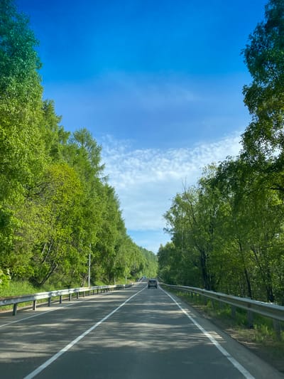 The image shows a view from a vehicle traveling on a paved road lined with guardrails and dense green trees on both sides. The road has white lane markings. The sky is bright blue with scattered white clouds. In the distance, a dark-colored car is visible on the road. The scene appears to be a natural setting, possibly a highway or a scenic route. There is no indication of the specific location within the city of Listvyanka, Irkutsk Oblast, Russia, as no landmarks or identifying features are present.