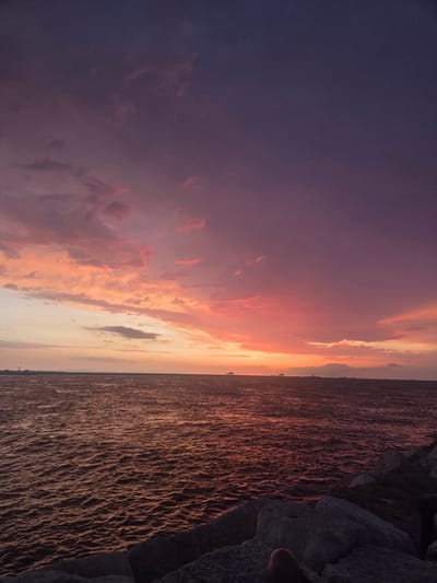 The image depicts a sunset over the Adriatic Sea, with the horizon visible and several structures and boats in the distance. The sky is filled with a dramatic display of color, transitioning from deep purple at the top to vibrant oranges and pinks closer to the horizon, indicating the sun has recently set or is about to set. Clouds are scattered across the sky, catching the light and contributing to the colorful spectacle. The sea reflects the colors of the sky, creating a shimmering, textured surface. In the foreground, large rocks are visible, suggesting a breakwater or shoreline. The scene is tranquil and evocative of a coastal sunset. While the image is of a coastal sunset, no specific landmarks or details definitively place it in Chioggia, Italy.