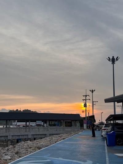 The image depicts a sunset scene in Pak Nam Chumphon, Thailand. The sky is predominantly gray with a vertical streak of orange and yellow light emanating from the setting sun. Utility poles and lighting fixtures are silhouetted against the sky. Along the right side of the image, there is a walkway with a blue painted surface and white markings. A person is standing on the walkway near parked cars and a blue trash bin. Below the walkway is a concrete wall or embankment with rocky terrain at its base. Further back, there are buildings and a bus or van parked under a covered structure.