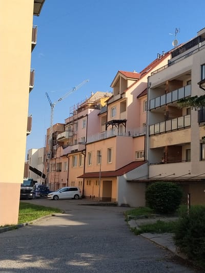 In Hluboká nad Vltavou, Czechia, on a sunny day, a street scene unfolds with residential buildings and construction activity. A tall construction crane is prominently positioned, its jib extending over a multi-story apartment complex that is undergoing renovations or expansion, indicated by scaffolding and exposed building materials. A white minivan is parked on the asphalted road in front of the buildings. Adjacent to the road, a strip of grass runs along a curb. To the left, a portion of a yellow-hued building with balconies is visible, partially obscuring the view of the construction site. The sky is clear and blue.
