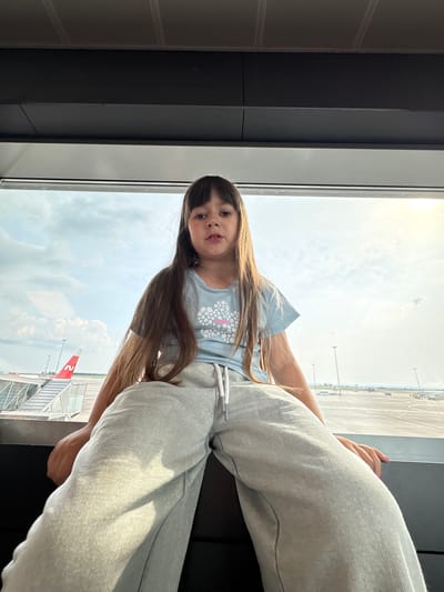 A low-angle, medium shot shows a young girl with long, dark brown hair sitting on a dark surface, looking directly at the camera. She is wearing a light blue t-shirt with a white floral design and light gray sweatpants. The girl is positioned in front of a large window, through which an airport tarmac is visible. An airplane with a red and white tail is parked to the left, with a jet bridge extended towards it. In the background, several other aircraft and airport vehicles are scattered across the tarmac, with lampposts lining the area. The sky is bright and partly cloudy, with the sun casting a warm glow. The scene appears to be a waiting area at an airport in Bol'shie Kabany, Russia.