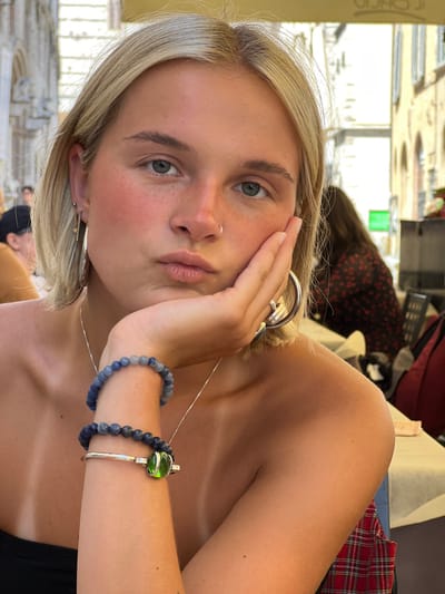 A fair-skinned woman with short blonde hair gazes towards the camera. She has a nose ring and wears large hoop earrings. Her left hand is placed on her cheek. She has a silver necklace, and two bracelets are visible on her right wrist. One bracelet is beaded and blue, while the other is silver with a green stone. The background is blurred, showing hints of an interior setting with other individuals. She is wearing a black top and appears to be seated.