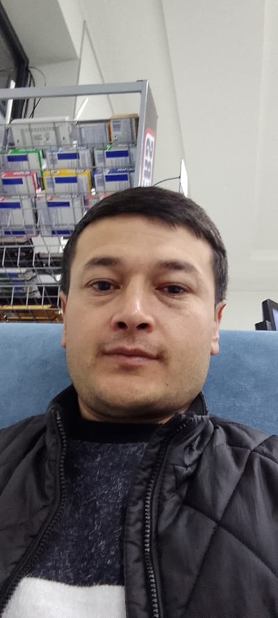 A man takes a selfie indoors, presumably in a shop, sitting on a blue cushioned bench. A black quilted jacket is visible over a patterned sweater, with a white shirt or undergarment partially showing. Behind the man is a retail display rack stocked with various boxed items and the upper corner of a computer monitor. The ceiling appears to be white with fluorescent lighting.