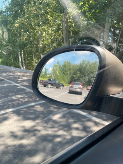 The image is taken from the perspective of a car interior, looking out through the passenger side window and reflecting in the side mirror. The scene captures a parking lot with trees in the background. In the reflection of the side mirror, a dark pickup truck is visible on the left, followed by a grey hatchback with its rear facing the camera. Further in the distance, a red car is also present. The parking lot pavement is marked with white lines and shows dappled sunlight patterns from the surrounding trees. The environment appears to be outdoors, likely in a suburban or rural setting, given the presence of mature trees and the absence of dense urban structures. The location is identified as Ivankovychi, Ukraine.