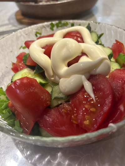 In Ozeretskoe, Russia, a close-up shot captures a bowl of salad. The salad is composed of chopped tomatoes and cucumbers, generously topped with a swirl of mayonnaise. The bowl, made of clear glass with a textured rim, sits on a white tablecloth that features a subtle floral pattern. In the background, slightly out of focus, is a glimpse of a dish with food and what appears to be a wooden placemat. The lighting suggests an indoor setting, possibly a home or restaurant.