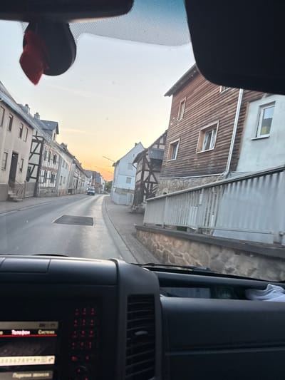 The image depicts a view from inside a vehicle, looking out at a street in Oberwetz, Germany. The perspective suggests the photo was taken by a passenger or driver looking forward and slightly upwards.
The scene features a narrow street lined with traditional German houses, characterized by their timber-framed facades and tiled roofs. Some of the houses appear to be older, while others show more modern renovations. The street itself is paved, with some markings visible, suggesting it's a residential area. In the distance, a white vehicle can be seen driving down the street, and the road curves slightly out of view.
The sky is bright and filled with a warm, golden light, indicating that it is either sunrise or sunset. The weather appears clear and calm.
Notable details include a black object mounted on the windshield, possibly a dashcam or GPS device, with a red cloth attached to it. Inside the vehicle, part of the dashboard is visible, including a digital display with red illuminated buttons and some text that is partially obscured but appears to be in Russian, with words like "Телефон" (Telephone) and "Поиск" (Search) discernible. The interior of the vehicle is dark, contrasting with the bright exterior.
The overall impression is a peaceful, everyday scene in a picturesque German town during a beautiful time of day.